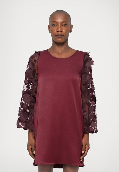 Femme portant une robe bordeaux avec des manches longues en appliqué floral transparent, se tenant devant un fond blanc uni.