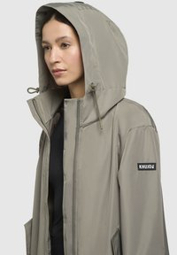 Lagani sivi softshell jakna s kapuljačom, prednjim zatvaračem i logotipom. Ima glatku teksturu i dva bočna džepa.