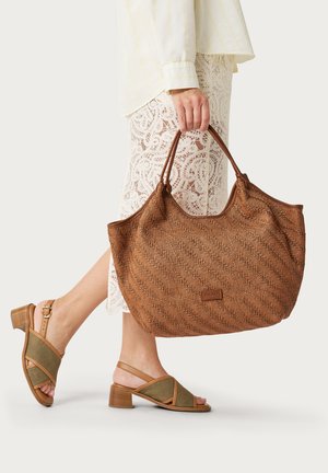 Personne portant une jupe en dentelle blanche et des sandales beige, tenant un grand sac cabas marron texturé avec doubles anses.