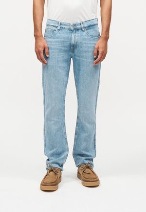 Moški v svetlo modrih kavbojkah s poševnim krojem, beli srajci in rjavih semiš čevljih z vezalkami, stoji na belem ozadju.