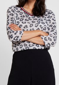 Haut à imprimé léopard en gris clair avec motifs noirs, col en V avec rayures rouges et noires, manches trois-quarts et coupe décontractée.