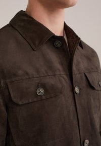 Chaqueta de ante marrón con cuello, que cuenta con dos bolsillos en el pecho y botones marrones. Textura suave con un corte entallado.