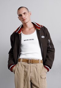 Brūna jakas ar sarkanu un baltu svītru apkakli, balta topiņš ar "Moschino" tekstu un bēšas krāsas bikses ar sānu kabatām un jostu.