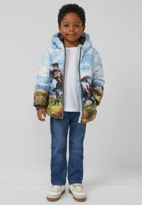 Gepolsterte Jacke mit Motiv eines blauen Himmels und Dinosauriern, ausgestattet mit einer Kapuze, Frontreißverschluss und elastischen Bündchen. Getragen mit einem weißen T-Shirt und blauen Jeans.