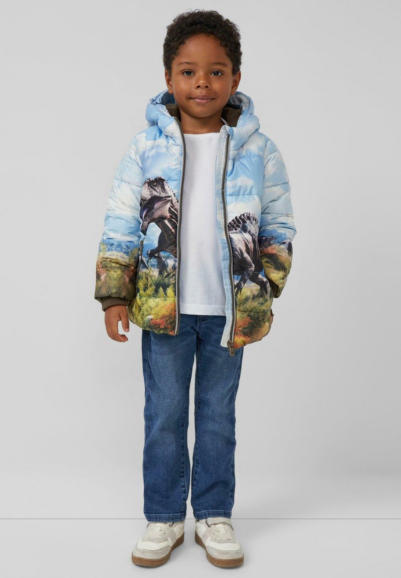 Gepolsterte Jacke mit Motiv eines blauen Himmels und Dinosauriern, ausgestattet mit einer Kapuze, Frontreißverschluss und elastischen Bündchen. Getragen mit einem weißen T-Shirt und blauen Jeans.