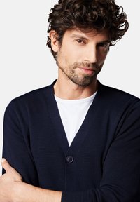 Granatowy kardigan wełniany z głębokim dekoltem w kształcie V i pojedynczym zapięciem na guzik. Noszony na białej koszulce, wyposażony w ściągacze przy mankietach i miękką fakturę.