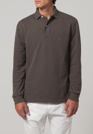 Polo à manches longues en coton brun foncé, avec un col et un petit logo sur la poitrine. Associé à un pantalon blanc.