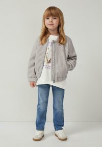 Junges Mädchen mit glattem, blonden Haar, das eine graue Kapuzenjacke, ein Sweatshirt mit Pferdedruck in Weiß, blaue Jeans und weiße Sneaker trägt und steht.