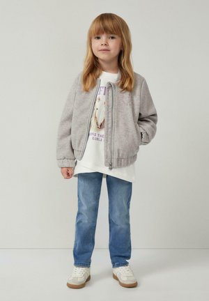 Junges Mädchen mit glattem, blonden Haar, das eine graue Kapuzenjacke, ein Sweatshirt mit Pferdedruck in Weiß, blaue Jeans und weiße Sneaker trägt und steht.