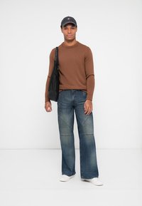 Pull en tricot marron, jean bleu à jambes larges avec des détails de couture, baskets blanches et casquette noire. Le modèle porte un sac noir sur une épaule.