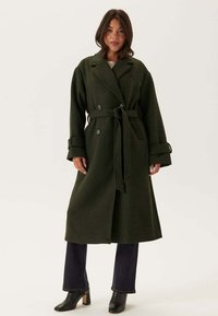 Cappotto lungo doppiopetto in lana verde scuro, con vita regolabile, rever oversize e polsini larghi. Abbinato a jeans scuri e stivali con tacco.