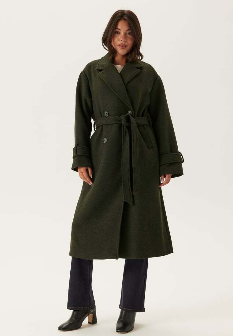 Cappotto lungo doppiopetto in lana verde scuro, con vita regolabile, rever oversize e polsini larghi. Abbinato a jeans scuri e stivali con tacco.