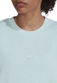 Sweat-shirt bleu clair en tissu doux, avec un col côtelé et un logo adidas blanc en relief sur la poitrine.
