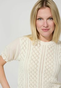 Cremfarbener Oberteil mit einem strukturierten Strickdesign und Rautenmuster sowie transparenten, puffigen Ärmeln. Der Stoff hat ein zartes und luftiges Aussehen.