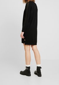 Abito nero a maglia con dettagli a coste sulle maniche, orlo spaccato e un piccolo logo. Abbinato a stivaletti neri e calzini bianchi.