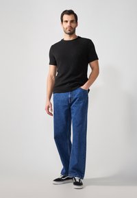 Camicetta nera a coste a maniche corte, jeans in denim blu dal taglio rilassato, e sneakers nere. Il modello si trova davanti a uno sfondo neutro.