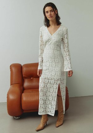 Hvid blondekjole med lange ærmer, V-udskæring og frontslids, kombineret med beige støvler med hæl. Baggrunden indeholder en brun lædersofa.