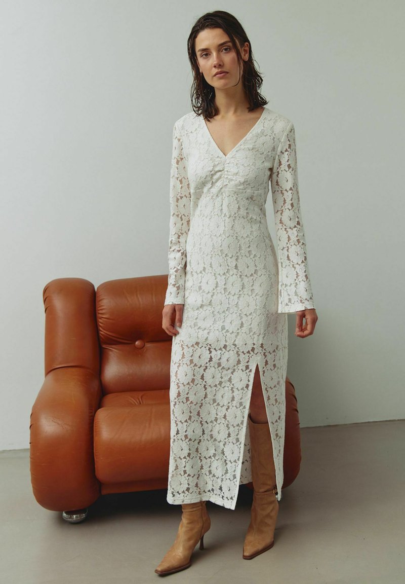 Valge pitskleit pikkade varrukatega, V-kaelusega ja ees lõhikuga, kombineeritud beežide kontsakingadega. Taustal on pruun nahast diivan.