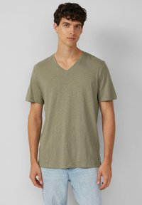 Młody mężczyzna z kręconymi włosami, ubrany w jasnooliwkowy T-shirt w serek i jasnobłękitne dżinsy, na jednolitym szarym tle.