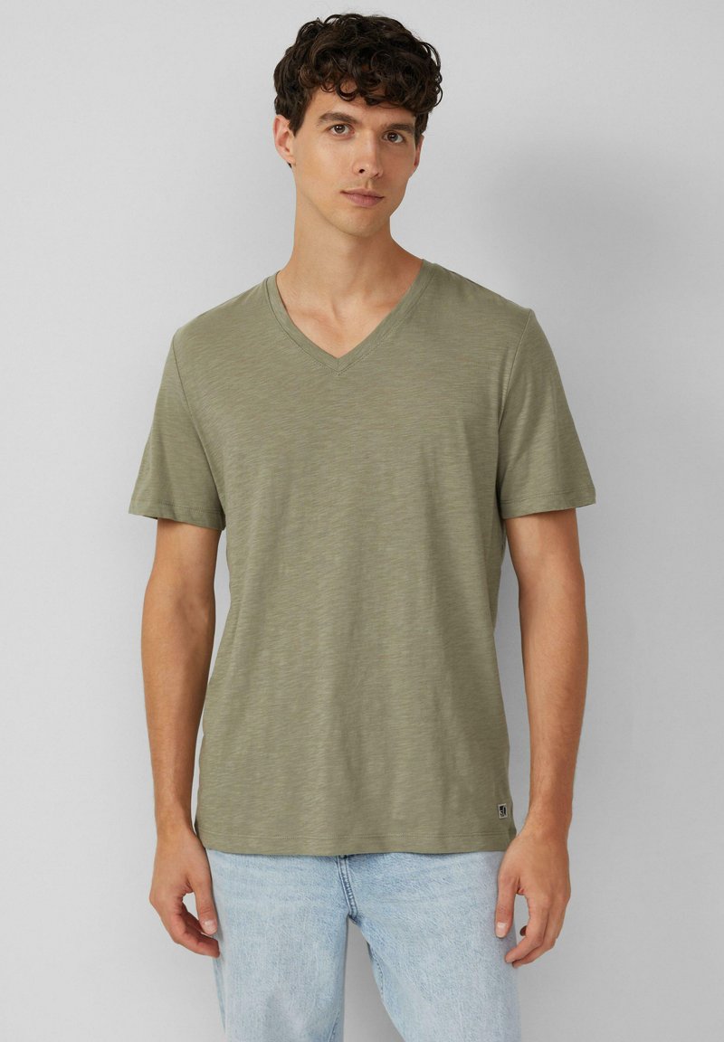 Młody mężczyzna z kręconymi włosami, ubrany w jasnooliwkowy T-shirt w serek i jasnobłękitne dżinsy, na jednolitym szarym tle.