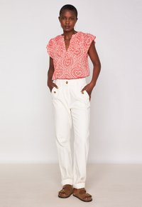 Blusa rossa a maniche corte con motivo, con scollatura a V, abbinata a pantaloni bianchi a vita alta con due tasche frontali e bottoni dorati.