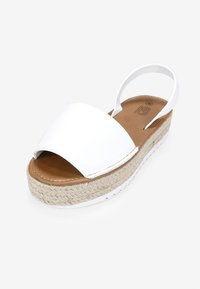 Sandalia de estilo espadrille blanca con una amplia banda delantera, plantilla marrón y plataforma de yute trenzado. Cuenta con una correa en la parte trasera para mayor soporte.