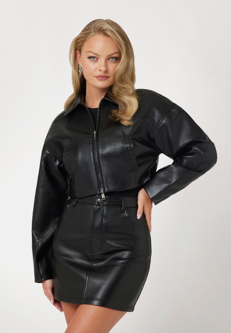 Juoda odinė trumpa striukė su priekiniu užtrauktuku, perteklinėmis rankovėmis ir juodu odiniu mini sijonu. Lygus paviršius ir minimalus detalizavimas.