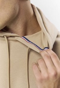 Beež kapuutsiga dressipluus, millel on nöör ja kontrastne sisemine kaelus, kus on punaseid ja siniseid triibusid. Pehme, sile tekstuur on nähtav.