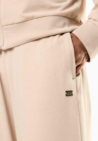 Chándal beige con una textura suave, que presenta un bolsillo visible y una etiqueta metálica con el logo en el lateral. Diseño simple y casual.