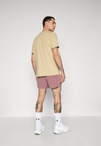 Beige T-Shirt, kurze rosa Shorts, weiße Sneaker und Sneakersocken. Siehe Tattoos an Armen und Beinen; schlichtes Design, keine Muster oder Akzente.