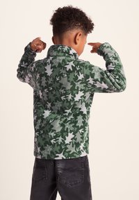 Green fleece pullover with a leaf pattern in various shades of green and gray. Features a high collar and long sleeves. Dark jeans.
