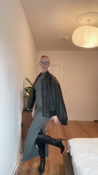 Juodas odinis švarkas, pilka suknelė, juodos aukštakulnės botai. Lygus audinys, elegantiškas dizainas. Vidinė aplinka su medinėmis grindimis ir augalu fone.