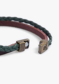 Pulsera trenzada de cuero verde con una base de cuero rojo oscuro. Presenta acentos de metal bronce con detalles grabados y un mecanismo de cierre.