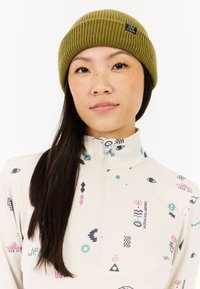 Olivovo zelený pletený čiapka s ribovaným vzorom a prišitým logom, nosená s vzorovaným, svetlo béžovým svetrom na zips, ktorý obsahuje abstraktné symboly v rôznych farbách.