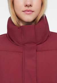 Chaqueta acolchada burdeos con un alto cuello, textura acolchada y detalles cosidos, diseñada para proporcionar calidez y comodidad.