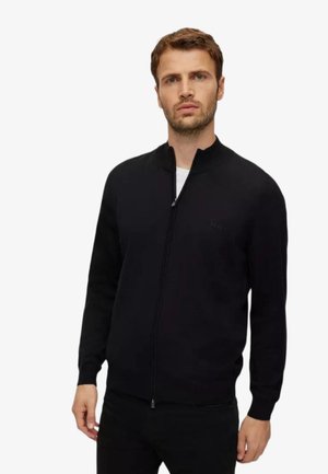 Uomo con capelli castani corti che indossa un maglione nero con cerniera sopra una camicia bianca, in piedi contro uno sfondo bianco uniforme.