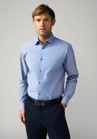 Světle modrá košile na knoflíky s jemným kostkovaným vzorem, strukturovaným límcem a dlouhými rukávy. V kombinaci s tmavými kalhotami a černým opaskem.