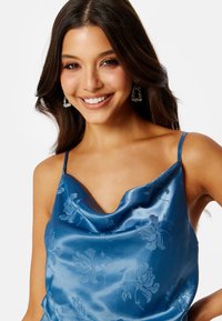 Niebieski satynowy top z dekoltem w serek, z wzorem w kwiaty i cienkimi ramiączkami. Modelka ma na sobie srebrne kolczyki w kształcie żyrandoli.