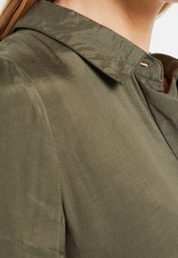 Oliwkowa bluzka o gładkiej fakturze, z klasycznym kołnierzykiem i detalami z guzikami w okolicy szyi. Lekko błyszczący materiał.