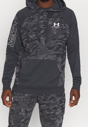 Mann trägt schwarzen und grauen Camouflage-Under Armour Hoodie und passende Hose, steht mit locker an den Seiten liegenden Händen.