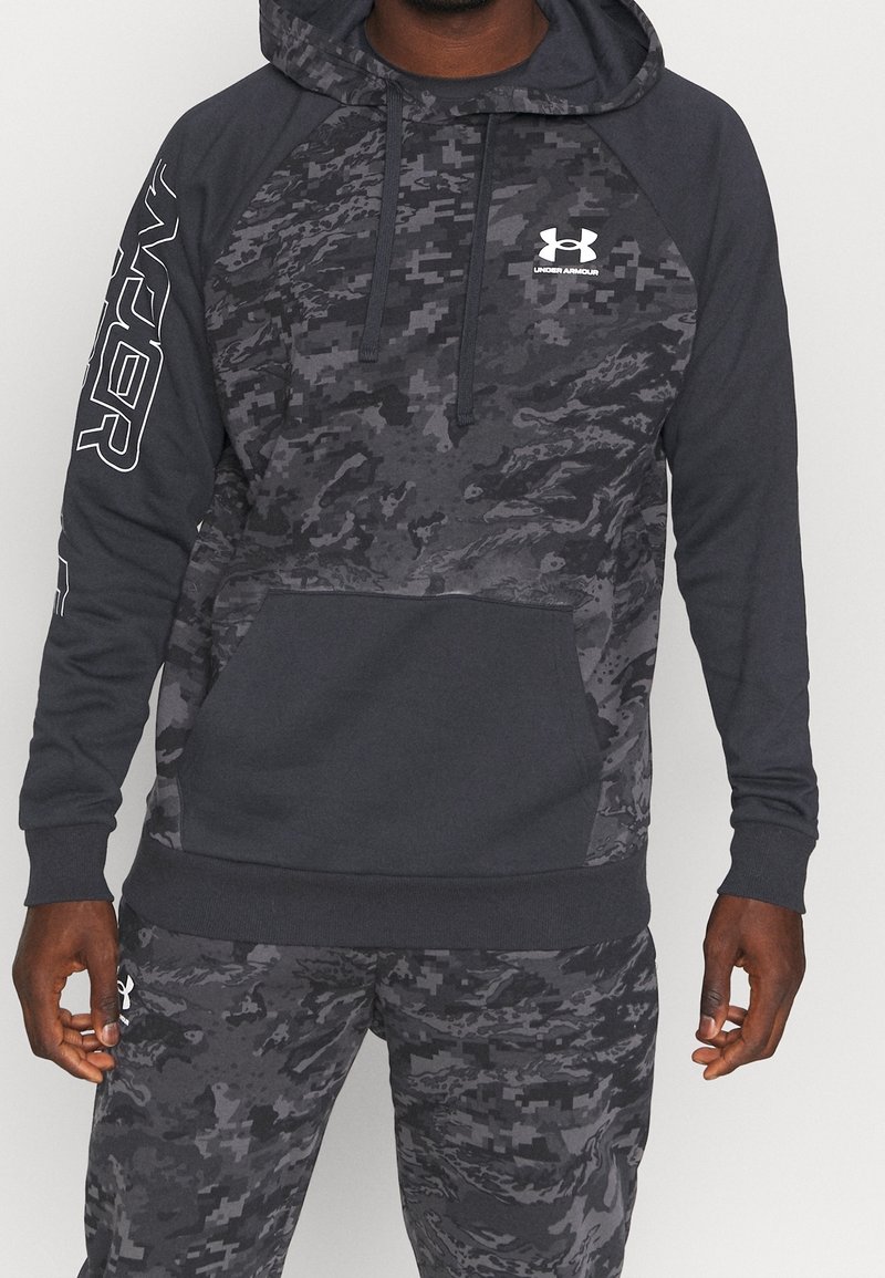 Homme portant un sweat à capuche Under Armour camouflage noir et gris avec un pantalon assorti, debout, les mains détendues le long du corps.