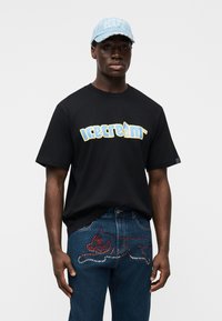 Μαύρο βαμβακερό t-shirt με το λογότυπο "icecream" σε κίτρινο και μπλε. Ανοιχτό μπλε σκουριασμένο καπέλο με κέντημα "HCT". Σκούρο μπλε τζιν με κόκκινο σχέδιο από στρας.