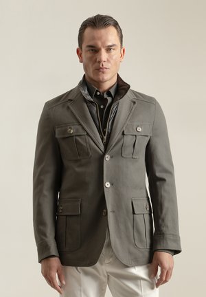 Chaqueta de mezcla de algodón gris con cuatro bolsillos de parche, cierre de botones y una capa interior negra desmontable. Estilizada con una camisa con cuello debajo.