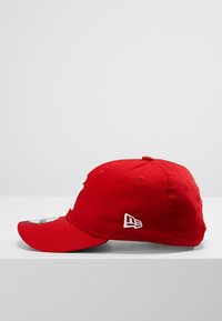 Boné de beisebol vermelho feito de algodão, com um aba curva, logo bordado na frente e uma pequena etiqueta branca na lateral.
