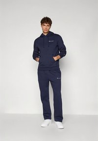 Námořnicky modrá mikina s kapucí, stahovacími šňůrkami a klokankovou kapsou, doplněná o odpovídající kalhoty; má decentní logo a volný střih.