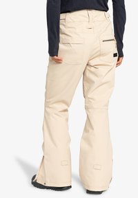 Beige snow pants with a relaxed fit, featuring multiple pockets, zippered rear pocket, and reinforced cuffs for durability.