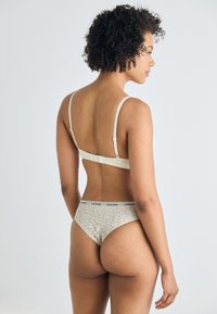 Calvin Klein undertøysett som inkluderer en beige BH med justerbare stropper og en matchende thong med mønstrete tekstur og logo-belte.