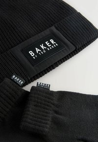 Schwarze Strickmütze und Handschuhe, beide mit dem Etikett "BAKER by Ted Baker", auf einem hellen Hintergrund präsentiert. Stylishe Winteraccessoires.
