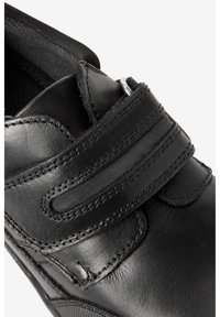 Chaussure en cuir noir avec une sangle auto-agrippante, des détails cousus et une texture lisse et brillante. La vue en gros plan met en évidence son design.
