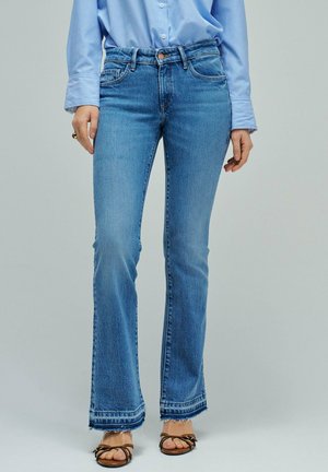 Femme portant une chemise bleu clair boutonnée et un jean évasé délavé moyen avec des ourlets effilochés et des sandales ouvertes, debout sur un fond uni.